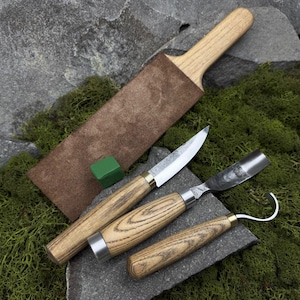 May include: A set of wood carving tools, including a knife, gouge, and hook knife, with light wood handles and metal blades. A leather strop and green sharpening stone are also included. The tools are displayed on a bed of green moss and gray stone.