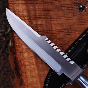 May include: A stainless steel survival knife with a serrated edge and a leather sheath. The knife blade is silver and the handle is a darker metal. The knife is on a wooden surface.