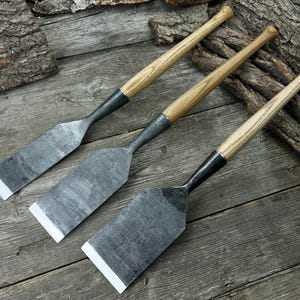 May include: Three hand-forged metal chisels with wooden handles. The blades are rectangular with a white edge. The handles are light-colored wood, and the metal is dark gray. The tools are arranged diagonally on a weathered wooden surface.