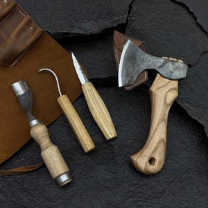 Puede incluir: Una colección de herramientas para tallar madera, que incluye un cincel, un cuchillo de gancho, un cuchillo recto y un hacha pequeña. Las herramientas tienen mangos de madera y hojas de metal. También se ve una funda de cuero y un rollo de herramientas.