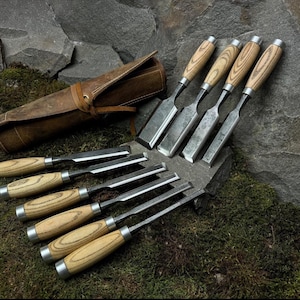 May include: A set of wood chisels with wooden handles and metal blades, arranged on a stone surface. A brown leather tool roll is also present. The chisels are of varying sizes, suitable for woodworking and carving projects.