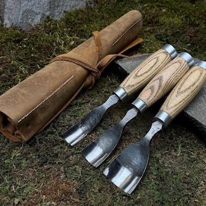 Może przedstawiać: Zestaw trzech dłut do rzeźbienia w drewnie z jasnymi drewnianymi uchwytami i metalowymi ostrzami, obok zwiniętego brązowego skórzanego wałka na narzędzia. Dłuta ułożone są na podłożu z zielonego mchu.