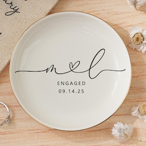 Op de afbeelding: Een witte keramische ringschaal met een gouden rand, met het woord 'engaged' en de datum 09.14.25 in zwart schrift. Een zilveren verlovingsring en een kalender liggen naast de schaal. Gedroogde bloemen zijn verspreid.