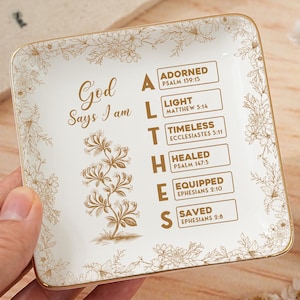 Puede incluir: Una bandeja cuadrada de cerámica blanca con borde dorado y detalles florales. La bandeja presenta el texto "Dios dice que soy" y una lista de palabras: Adornado, Luz, Atemporal, Sanado, Equipado y Salvado, con versículos bíblicos.