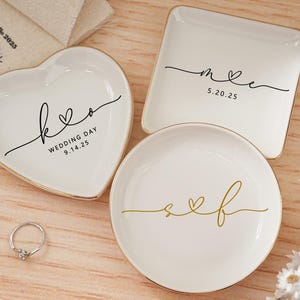 May include: White ceramic ring dishes in heart, square, and round shapes, with gold rims. The heart-shaped dish has the text "k♡o WEDDING DAY 9.14.25". The square dish has "m♡e 5.20.25". The round dish has "s♡f". A silver ring is also present.