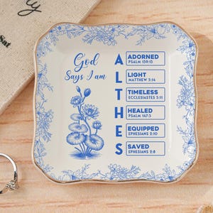 Puede incluir: Un plato cuadrado de cerámica blanca con un diseño floral azul y ribete dorado. El plato presenta el texto "Dios dice que soy" y una lista de afirmaciones. Un anillo y flores decorativas están en el fondo.