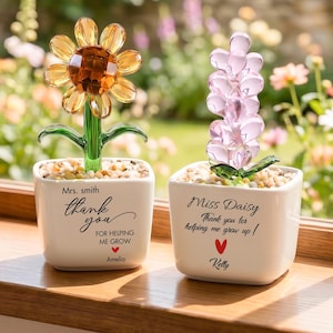 May include: Two white ceramic square planters containing glass flowers. The left planter features a yellow sunflower and text reading Mrs. smith thank you FOR HELPING ME GROW Amelia. The right planter has a pink flower and text reading Miss Daisy Thank you for helping me grow up ! Kelly.