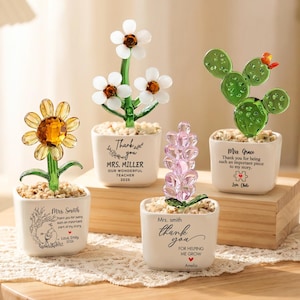 Puede incluir: Cuatro adornos de plantas de cristal en macetas de cerámica cuadradas blancas rellenas de piedras pequeñas. Las plantas incluyen un girasol amarillo, una flor blanca, una flor rosa y un cactus verde. Cada maceta tiene texto personalizado.