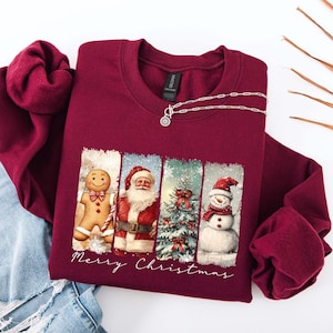 Puede incluir: Sudadera burdeos de cuello redondo con un dise&ntilde;o festivo. El gr&aacute;fico presenta un hombre de jengibre, Pap&aacute; Noel, un &aacute;rbol de Navidad y un mu&ntilde;eco de nieve. Las palabras "Merry Christmas" est&aacute;n escritas debajo. Un collar plateado est&aacute; sobre la sudadera.