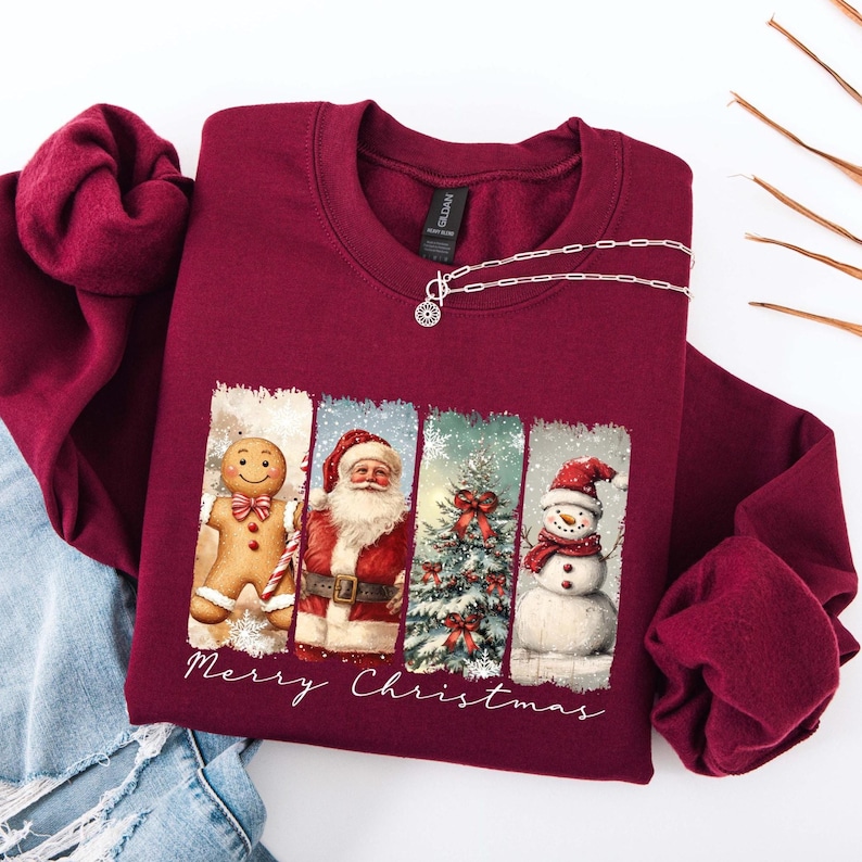 Puede incluir: Sudadera burdeos de cuello redondo con un dise&ntilde;o festivo. El gr&aacute;fico presenta un hombre de jengibre, Pap&aacute; Noel, un &aacute;rbol de Navidad y un mu&ntilde;eco de nieve. Las palabras "Merry Christmas" est&aacute;n escritas debajo. Un collar plateado est&aacute; sobre la sudadera.