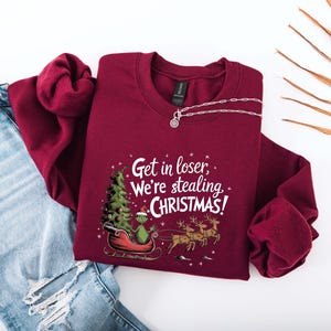 May include: Burgundy crewneck sweatshirt with the text "Get in loser, We're stealing CHRISTMAS!". The graphic features a green character in a sleigh with a Christmas tree and reindeer. A silver necklace is also visible.