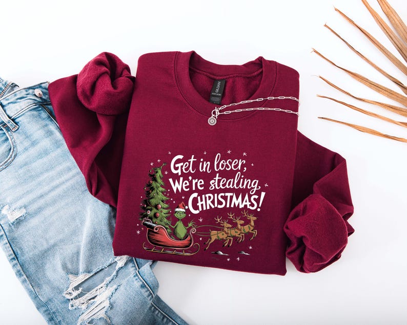 May include: Burgundy crewneck sweatshirt with the text "Get in loser, We're stealing CHRISTMAS!". The graphic features a green character in a sleigh with a Christmas tree and reindeer. A silver necklace is also visible.