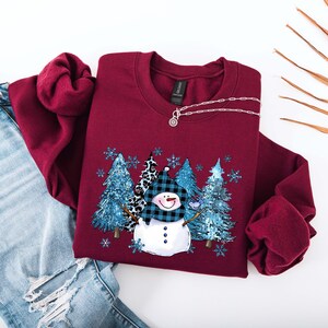 May include: Burgundy crewneck sweatshirt featuring a snowman design with a blue plaid hat and a forest scene. The design includes blue trees and snowflakes. A silver necklace is draped on the sweatshirt. The sweatshirt is paired with blue jeans.