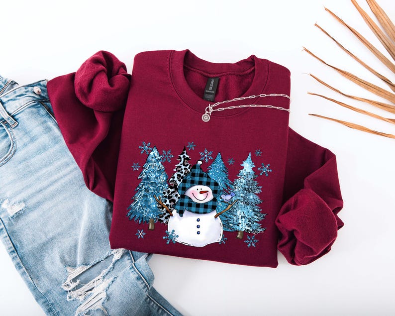 May include: Burgundy crewneck sweatshirt featuring a snowman design with a blue plaid hat and a forest scene. The design includes blue trees and snowflakes. A silver necklace is draped on the sweatshirt. The sweatshirt is paired with blue jeans.