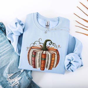 May include: A light blue sweatshirt featuring a pumpkin design made of books. The pumpkin has a green stem and is surrounded by curly vines. The books are various colors, including orange, red, and brown. The sweatshirt is paired with light blue jeans.