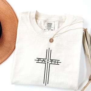 May include: Cream-colored t-shirt with a black cross design and the word "FAITH" in the center. A gold necklace with a pendant rests on the shirt. The shirt is from the Comfort Colors brand.