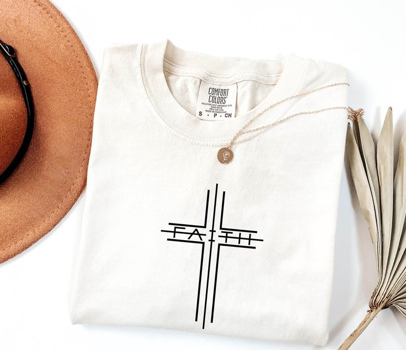 May include: Cream-colored t-shirt with a black cross design and the word "FAITH" in the center. A gold necklace with a pendant rests on the shirt. The shirt is from the Comfort Colors brand.