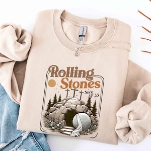 May include: Beige sweatshirt featuring the text "Rolling Stones Since AD 33" above an illustration of a tomb with three crosses, trees, and a large stone. A silver necklace with a sun pendant is on the sweatshirt.