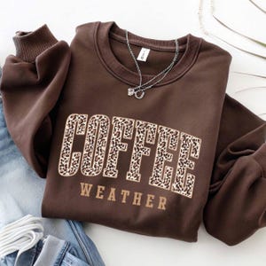 May include: A brown sweatshirt with the words "COFFEE WEATHER" in a leopard print design. The sweatshirt has a crew neck and long sleeves. A silver necklace with a small pendant is draped on the sweatshirt.