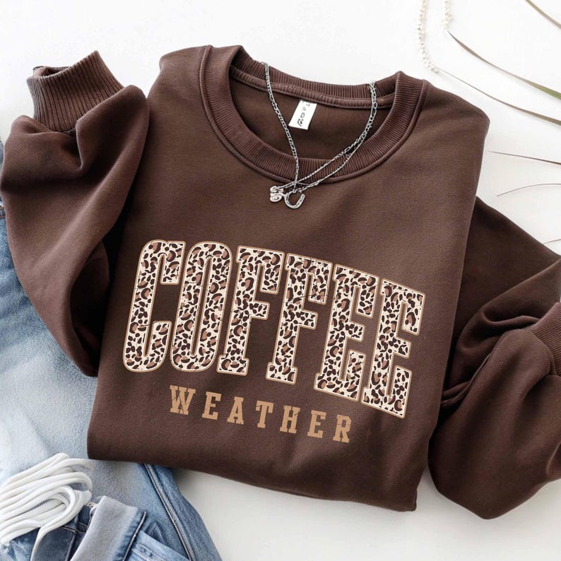 May include: A brown sweatshirt with the words "COFFEE WEATHER" in a leopard print design. The sweatshirt has a crew neck and long sleeves. A silver necklace with a small pendant is draped on the sweatshirt.