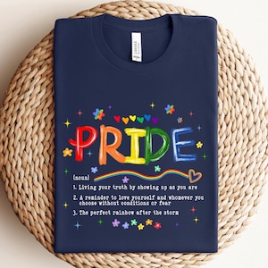 May include: Navy blue t-shirt with the word "PRIDE" in rainbow colors, surrounded by colorful hearts, stars, and flowers. The shirt includes a definition of pride: "Living your truth by showing up as you are."