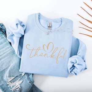 May include: A light blue sweatshirt with the word "thankful" in gold script and a heart design. The sweatshirt is folded next to a pair of blue jeans with distressed details. A silver necklace is draped across the top of the sweatshirt.