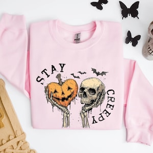 May include: A light pink sweatshirt with a Halloween-themed graphic. The graphic features a jack-o'-lantern heart and a skull held by skeletal hands, with the words "STAY CREEPY" in black. Black bats and butterflies are scattered around.