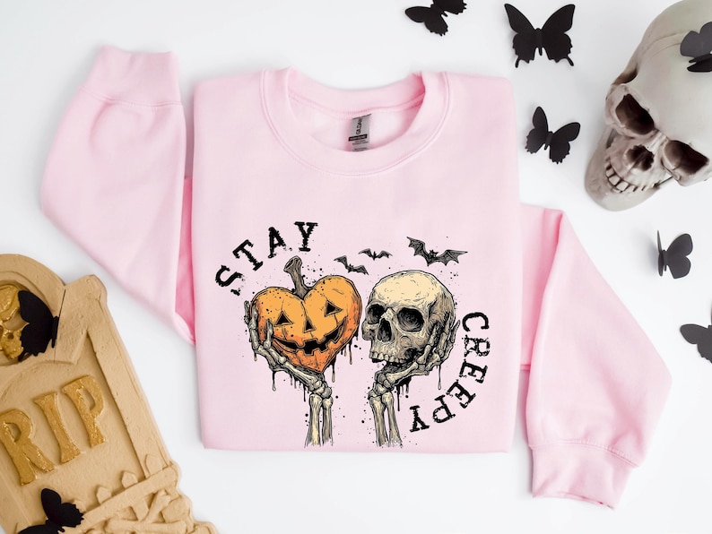 May include: A light pink sweatshirt with a Halloween-themed graphic. The graphic features a jack-o'-lantern heart and a skull held by skeletal hands, with the words "STAY CREEPY" in black. Black bats and butterflies are scattered around.
