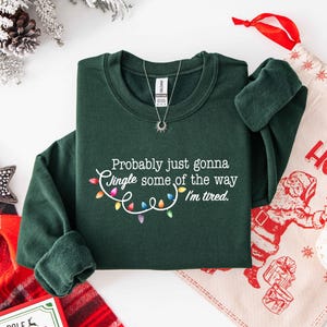 Puede incluir: Una sudadera verde oscuro con el texto "Probably just gonna jingle some of the way I'm tired." El texto est&aacute; acompa&ntilde;ado de una cadena de luces navide&ntilde;as de colores. Se ve un collar plateado con un colgante de sol.