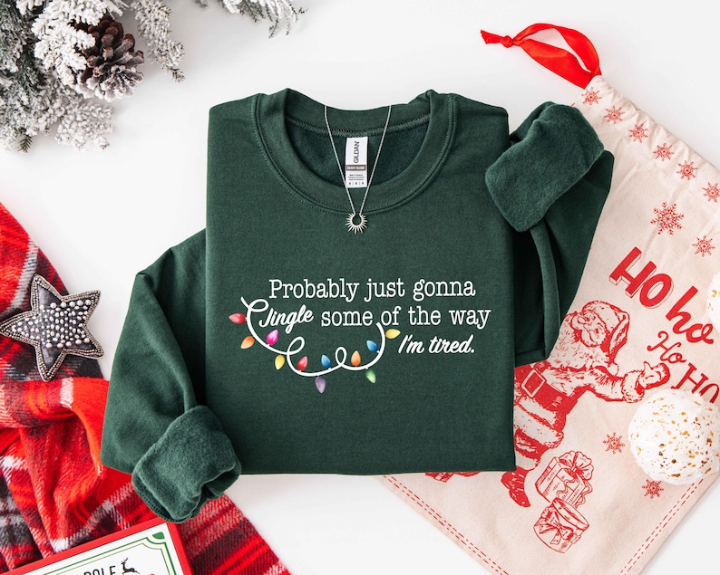 Puede incluir: Una sudadera verde oscuro con el texto "Probably just gonna jingle some of the way I'm tired." El texto est&aacute; acompa&ntilde;ado de una cadena de luces navide&ntilde;as de colores. Se ve un collar plateado con un colgante de sol.