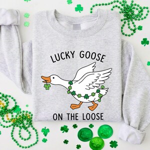 May include: A light gray sweatshirt with the text "LUCKY GOOSE ON THE LOOSE" and a cartoon goose wearing a shamrock garland. The goose is holding a shamrock in its beak. Green shamrock decorations and beads surround the sweatshirt.