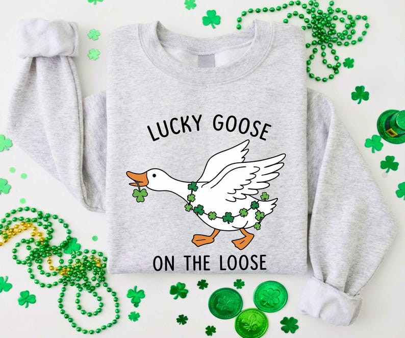 May include: A light gray sweatshirt with the text "LUCKY GOOSE ON THE LOOSE" and a cartoon goose wearing a shamrock garland. The goose is holding a shamrock in its beak. Green shamrock decorations and beads surround the sweatshirt.