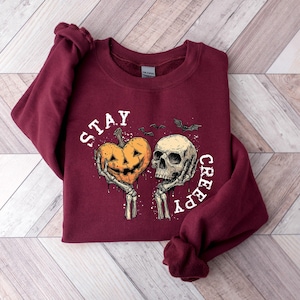 May include: Burgundy sweatshirt with a Halloween-themed graphic. The design features a jack-o'-lantern heart and a skull held by skeleton hands, with the words "STAY CREEPY" in white. Two bats are also included in the design.