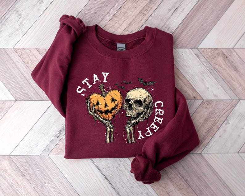 May include: Burgundy sweatshirt with a Halloween-themed graphic. The design features a jack-o'-lantern heart and a skull held by skeleton hands, with the words "STAY CREEPY" in white. Two bats are also included in the design.