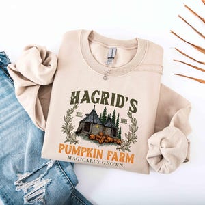 May include: Beige crewneck sweatshirt featuring a graphic of Hagrid's Pumpkin Farm. The design includes a house, pumpkins, and text that reads "Hagrid's Pumpkin Farm Magically Grown." The sweatshirt is paired with blue jeans.