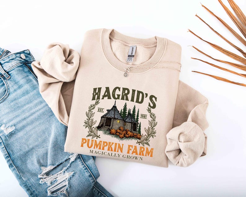 May include: Beige crewneck sweatshirt featuring a graphic of Hagrid's Pumpkin Farm. The design includes a house, pumpkins, and text that reads "Hagrid's Pumpkin Farm Magically Grown." The sweatshirt is paired with blue jeans.