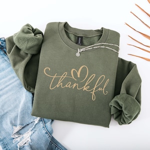 May include: An olive green sweatshirt with the word "thankful" in a cursive font and a heart design. The sweatshirt is paired with light blue ripped jeans and a silver necklace. A fall-themed fashion item.