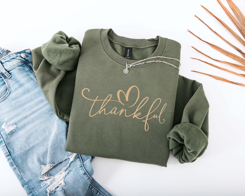 May include: An olive green sweatshirt with the word "thankful" in a cursive font and a heart design. The sweatshirt is paired with light blue ripped jeans and a silver necklace. A fall-themed fashion item.