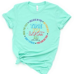 May include: A light turquoise t-shirt with a colorful graphic. The graphic reads "Time to Lock In" surrounded by phrases like "Rock the Test" and "You Can Do This." The shirt is knotted at the bottom.