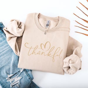 May include: A beige sweatshirt with the word "thankful" in a cursive font and a heart design. The sweatshirt is folded and placed next to a pair of blue jeans with distressed details. A silver necklace is visible on the sweatshirt.