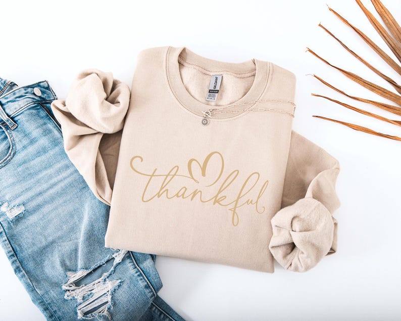 May include: A beige sweatshirt with the word "thankful" in a cursive font and a heart design. The sweatshirt is folded and placed next to a pair of blue jeans with distressed details. A silver necklace is visible on the sweatshirt.