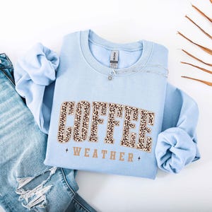 May include: A light blue sweatshirt with the words "COFFEE WEATHER" in a leopard print design. The sweatshirt is paired with light wash denim jeans. A silver necklace is draped across the sweatshirt.