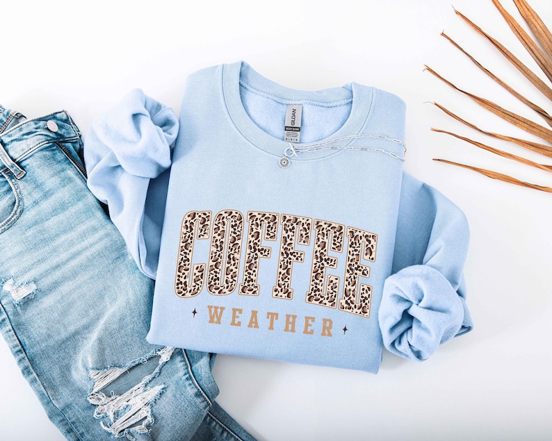 May include: A light blue sweatshirt with the words "COFFEE WEATHER" in a leopard print design. The sweatshirt is paired with light wash denim jeans. A silver necklace is draped across the sweatshirt.