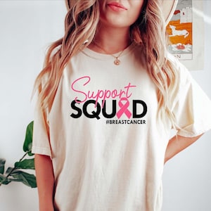 May include: Cream-colored t-shirt with the words "Support SQUAD" in pink and black, featuring a pink ribbon graphic. The hashtag "#BREASTCANCER" is printed below the text. The shirt is a casual style.