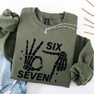 May include: Olive green sweatshirt with the words "SIX SEVEN" and a skeleton hand graphic. A silver necklace with a sun pendant is draped on the sweatshirt. The sweatshirt is folded on top of blue jeans.
