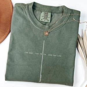 May include: An olive green t-shirt with the text "THE WAY, THE TRUTH, AND THE LIFE" and "JOHN 14:6" printed in white. A gold necklace with a small pendant rests on the shirt. The shirt is folded and has a tag that says "COMFORT COLORS".
