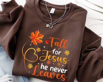 May include: A brown sweatshirt with the text "Fall for Jesus he never Leaves" in orange and white. The design includes fall leaves in orange and yellow. A silver necklace with a horseshoe pendant is on top of the sweatshirt.