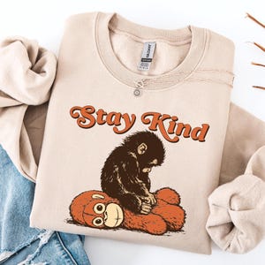 May include: A beige sweatshirt with the words "Stay Kind" in orange lettering. A brown illustration of a monkey is sitting on a brown teddy bear. A silver necklace with a sun pendant is on the sweatshirt.