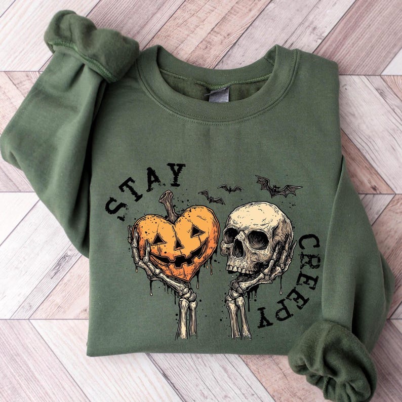 May include: Green sweatshirt with a Halloween-themed graphic. The design features a jack-o'-lantern heart and a skull held by skeleton hands, with the words "STAY CREEPY" and bats. The sweatshirt has long sleeves.