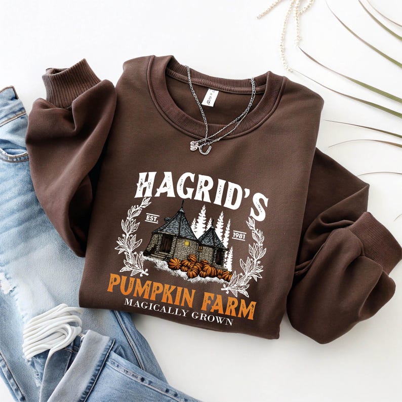 May include: A brown sweatshirt with the text "Hagrid's Pumpkin Farm" in white and orange, featuring a graphic of a building and pumpkins. The sweatshirt has long sleeves and a crew neck. A silver necklace is draped across the front.